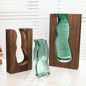 May include: Three decorative vases. One is a clear, textured, teal-coloured glass vase. Two are wooden frames with a carved-out shape, one holding a teal glass vase. The wood has a dark brown finish. The vases are on a white surface.