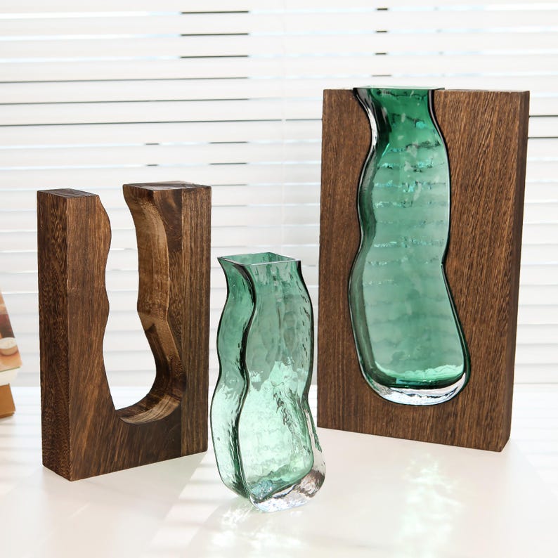 May include: Three decorative vases. One is a clear, textured, teal-coloured glass vase. Two are wooden frames with a carved-out shape, one holding a teal glass vase. The wood has a dark brown finish. The vases are on a white surface.