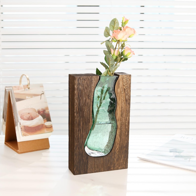 May include: A decorative vase with a clear, organically shaped glass insert set in a dark brown wooden frame. The vase holds a small bouquet of pink roses. The design is modern and artistic, suitable for home decor.