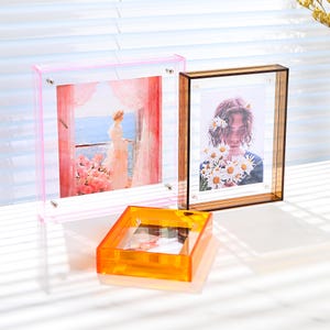 Pode incluir: Tr&ecirc;s molduras de fotos de acr&iacute;lico transparente em rosa, castanho e laranja. A moldura rosa cont&eacute;m uma foto de uma mulher olhando por uma janela. A moldura castanha tem uma foto de uma pessoa segurando flores. A moldura laranja &eacute; quadrada.