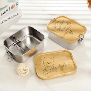 May include: Two stainless steel lunchboxes with bamboo lids. One lid features a cartoon elephant with the name "Alice" engraved on it. The other lid features a cartoon bear with the name "Jackson" engraved on it.