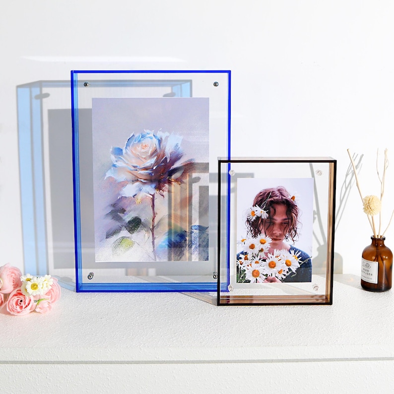 Pode incluir: Duas molduras de acr&iacute;lico, uma com borda azul e outra com borda preta, exibem obras de arte e uma fotografia. A moldura com borda azul cont&eacute;m uma impress&atilde;o floral, enquanto a moldura com borda preta apresenta uma pessoa segurando margaridas.