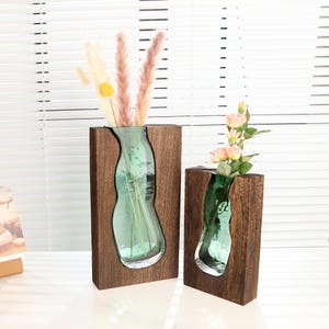 May include: Two decorative vases. Each vase features a clear, green glass insert set within a dark brown wooden frame. One vase holds dried flowers, while the other holds pink roses. The vases are designed to hold flowers.