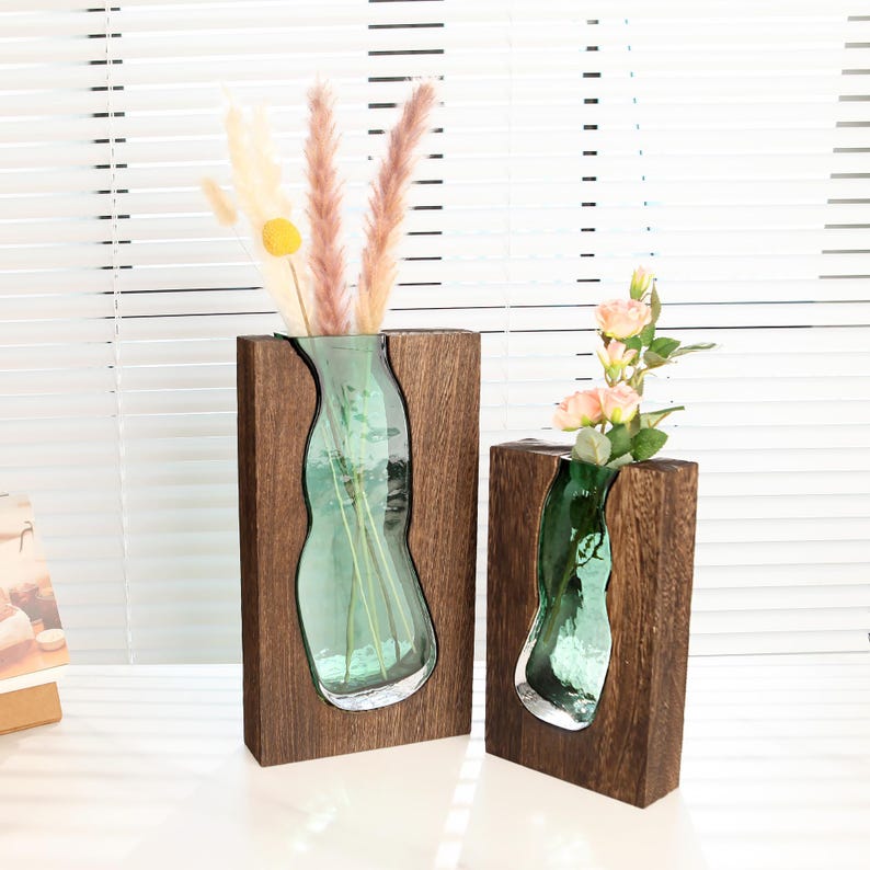 May include: Two decorative vases. Each vase features a clear, green glass insert set within a dark brown wooden frame. One vase holds dried flowers, while the other holds pink roses. The vases are designed to hold flowers.