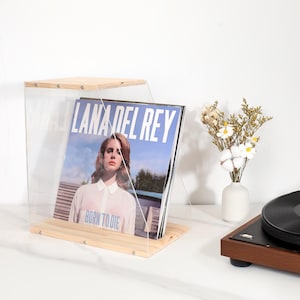 May include: A clear acrylic record display case with a wooden base and top. The case holds a vinyl record album with the text "LANA DEL REY BORN TO DIE". A white vase with flowers and a record player are also visible.