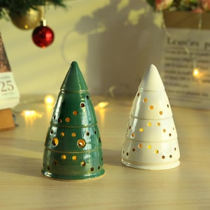 May include: Two ceramic Christmas tree decorations. One is a glossy green, the other is white. Both have a conical shape with circular cutouts. The trees are designed to hold a small light inside, creating a warm glow.
