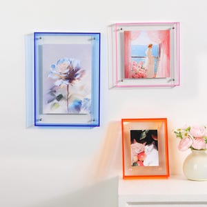 Pode incluir: Tr&ecirc;s molduras de fotos em acr&iacute;lico azul, rosa e laranja. A moldura azul cont&eacute;m uma impress&atilde;o floral, a moldura rosa apresenta uma cena mar&iacute;tima e a moldura laranja exibe uma foto de gato. As molduras s&atilde;o montadas em uma parede branca.