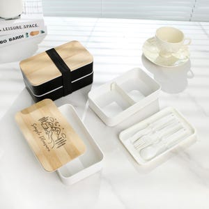 May include: A two-tiered wooden lunch box with a black base and a white lid. The lid has a bamboo top with the text "Simple Pleasure" and a camping scene. The lunch box comes with a white container with two compartments and a white tray with a fork, spoon, and chopsticks.