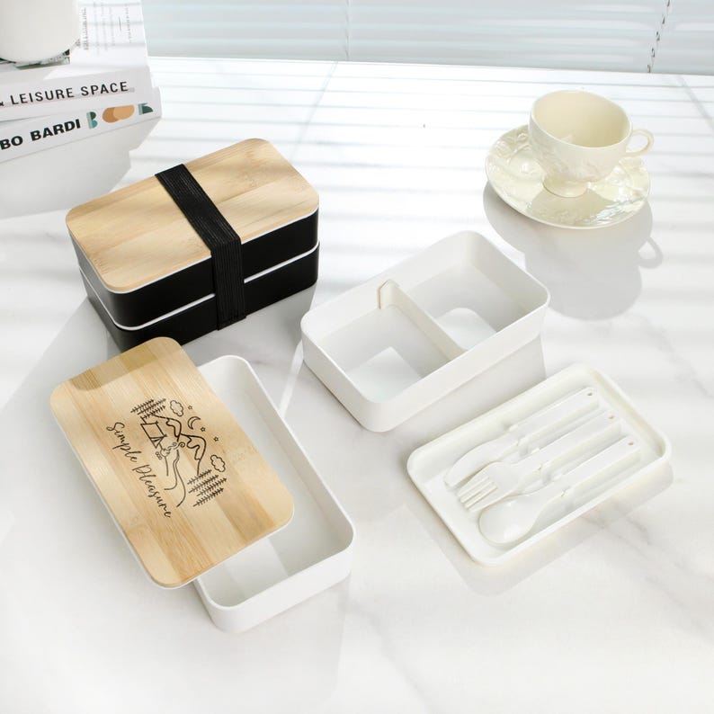 May include: A two-tiered wooden lunch box with a black bottom and a white lid. The lid has a bamboo top with the text "Simple Pleasure" and a camping scene. The lunch box comes with a white container with two compartments and a white tray with a fork, spoon, and chopsticks.
