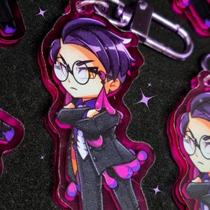 Peut inclure: Un porte-clés en acrylique violet et rose représentant un personnage de dessin animé aux cheveux violets, des lunettes et un costume noir. Le porte-clés a un fermoir argenté et est entouré d'une bordure rose. D'autres porte-clés sont visibles.