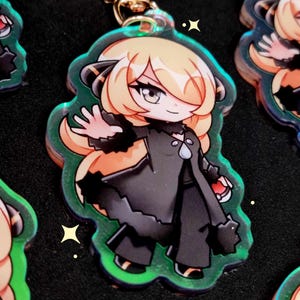 May include: An acrylic keyring featuring a chibi-style character with long blonde hair, wearing a black coat with a high collar and a silver teardrop pendant. The character has one hand raised and holds a small red and white sphere in the other.