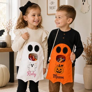 Può includere: Due sacchetti in tessuto per dolcetti con tracolla. Un sacchetto è bianco con un disegno di un fantasma e il nome Ruby, l'altro è arancione con un disegno di una zucca e il nome Mason. Entrambi presentano occhi da fantasma neri e un'apertura centrale.