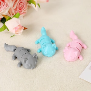 May include: Three cat-shaped silicone tea infusers in grey, blue, and pink. The infusers are segmented, allowing them to open and close. The cat's face and paws are visible. The background includes pink roses and a white surface.