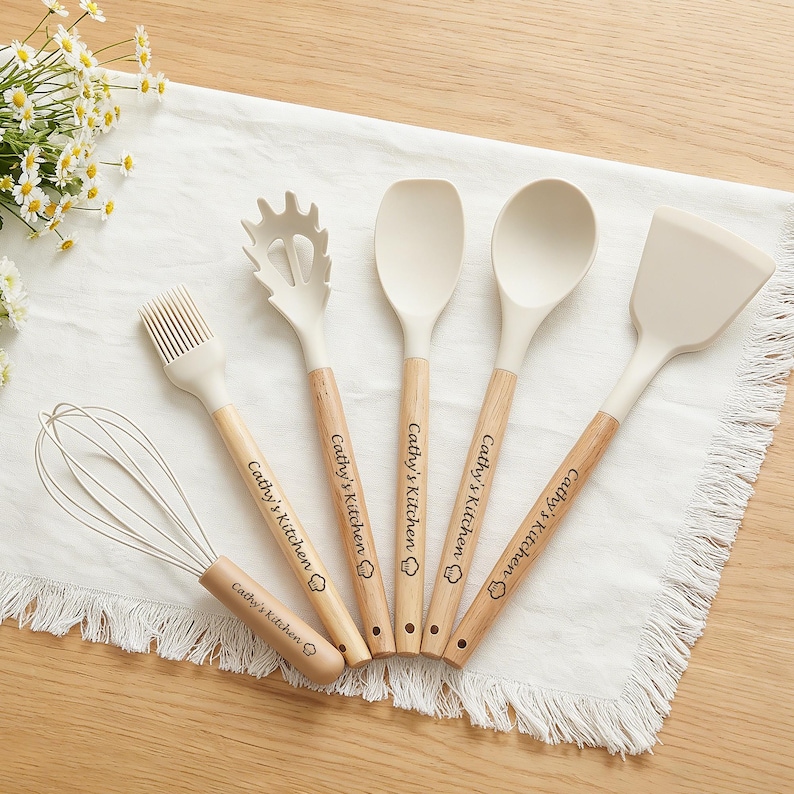 K&ouml;nnte beinhalten: Ein Set aus sechs K&uuml;chenutensilien, darunter ein Schneebesen, ein Pinsel, ein Spatel, ein L&ouml;ffel und eine Nudelkelle. Jedes Utensil hat einen hellbeigen Kopf und einen Holzgriff. Der Text "Cathy's Kitchen" ist auf jedem Griff aufgedruckt.
