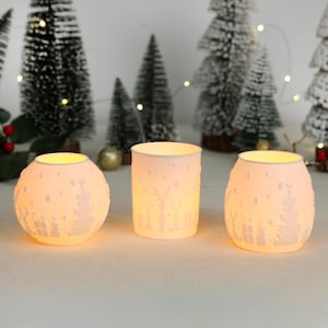 Op de afbeelding: Drie gloeiende witte keramische kandelaars met feestelijke uitgesneden ontwerpen. De kandelaars hebben de vorm van een bol, een cilinder en een afgeronde vorm. De uitgesneden ontwerpen omvatten sterren, bomen en rendieren. De achtergrond omvat miniatuur kerstbomen.