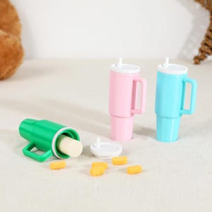 May include: A green, pink, and blue tumbler set with white lids and handles. The green tumbler is open, revealing a white cylindrical object. Several small yellow objects are scattered around the tumblers.