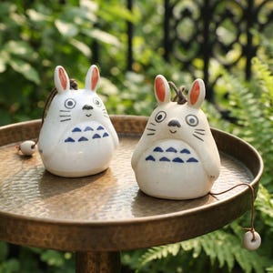 May include: Two white ceramic bunny figurines with orange-tipped ears, black eyes, and blue geometric patterns on their bellies. Each figurine has a string and a small, round bead attached. They are displayed on a bronze-colored tray.