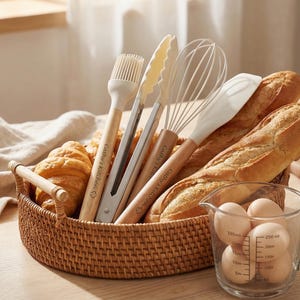 K&ouml;nnte beinhalten: Ein Weidenkorb gef&uuml;llt mit Backwaren, darunter Croissants und Baguettes, und Backutensilien. Die Utensilien umfassen einen Pinsel, eine Zange, einen Schneebesen und einen Spatel. Ein Messbecher mit Eiern ist ebenfalls vorhanden. Der Text "Cathy's Kitchen" steht auf den Werkzeugen.