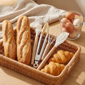 K&ouml;nnte beinhalten: Ein Weidenkorb enth&auml;lt zwei Baguettes, Croissants und Backutensilien. Die Werkzeuge umfassen einen Pinsel, eine Zange, einen Schneebesen und einen Spatel. Ein Pyrex-Messbecher mit Eiern befindet sich im Hintergrund. Die Werkzeuge tragen die Aufschrift "Cathy's Kitchen".