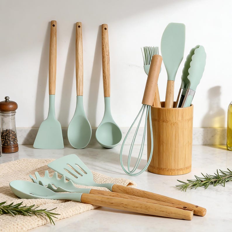 K&ouml;nnte beinhalten: Eine Sammlung von mintgr&uuml;nen K&uuml;chenutensilien mit Holzgriffen. Das Set umfasst einen Spatel, einen L&ouml;ffel, eine Kelle, einen Schneebesen, einen Pinsel, eine Zange und einen Bambus-Utensilienhalter. Die Utensilien sind auf einer wei&szlig;en Arbeitsplatte mit einem beigen Tuch angeordnet.