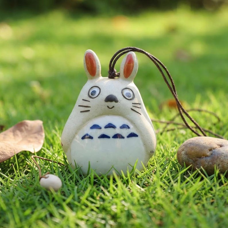 May include: A ceramic figurine of a cartoon rabbit with a brown cord for hanging. The rabbit is white with orange-tipped ears, blue markings on its belly, and black details. It is set against a backdrop of green grass.
