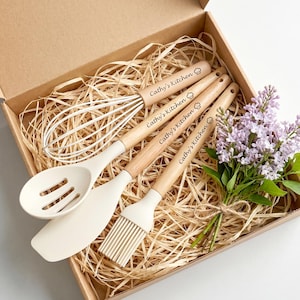 May include: A boxed set of kitchen utensils, including a whisk, slotted spoon, spatula, and basting brush. The wooden handles are engraved with "Cathy's Kitchen". The utensils are cream-colored and rest on a bed of straw. A bouquet of purple flowers is included.