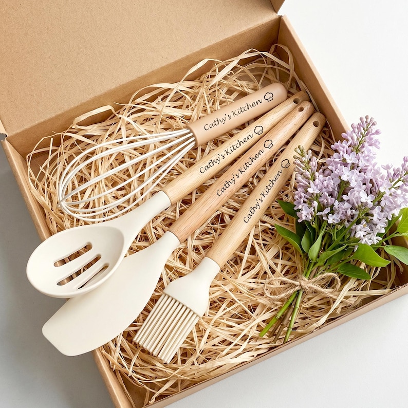 K&ouml;nnte beinhalten: Ein K&uuml;chenutensilien-Set in einer Box, einschlie&szlig;lich Schneebesen, Sch&ouml;pfl&ouml;ffel, Spatel und Backpinsel. Die Holzgriffe sind mit "Cathy's Kitchen" graviert. Die Utensilien sind cremefarben und liegen auf einem Strohbett. Ein Strau&szlig; lila Blumen ist enthalten.
