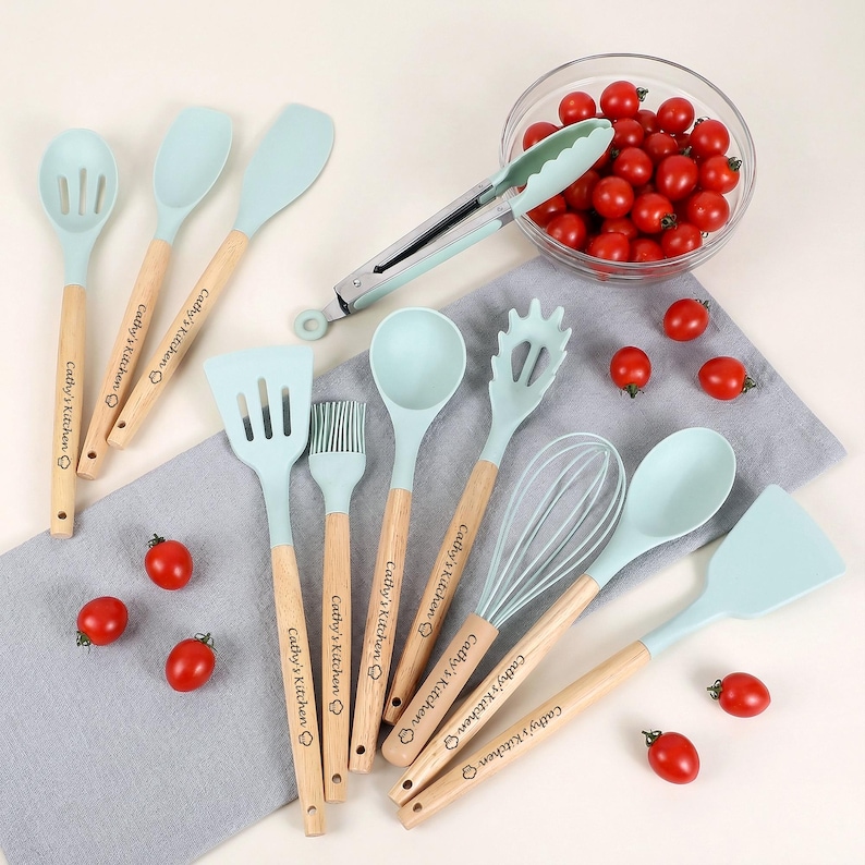 K&ouml;nnte beinhalten: Eine Sammlung hellblauer K&uuml;chenutensilien mit Holzgriffen, darunter Spatel, L&ouml;ffel, ein Schneebesen und eine Zange. Die Griffe sind mit "Cathy's Kitchen" graviert. Eine Sch&uuml;ssel mit roten Cherrytomaten ist ebenfalls vorhanden.
