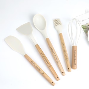 K&ouml;nnte beinhalten: Ein Set aus f&uuml;nf K&uuml;chenutensilien: ein Spatel, ein L&ouml;ffel, ein Pfannenwender, ein Schneebesen und ein Backpinsel. Jedes Utensil hat einen hellfarbigen Silikonkopf und einen Holzgriff. Der Text "Cathy's Kitchen" ist auf jedem Griff aufgedruckt.