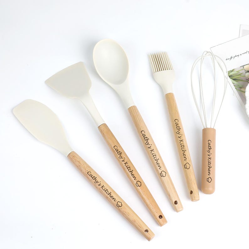 K&ouml;nnte beinhalten: Ein Set aus f&uuml;nf K&uuml;chenutensilien: ein Spatel, ein L&ouml;ffel, ein Pfannenwender, ein Schneebesen und ein Backpinsel. Jedes Utensil hat einen hellfarbigen Silikonkopf und einen Holzgriff. Der Text "Cathy's Kitchen" ist auf jedem Griff aufgedruckt.
