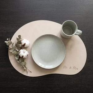 Puede incluir: Un mantel individual beige con un plato a rayas verde claro, una taza gris y una ramita de algodón y eucalipto. Las palabras "Mr & Mrs" están impresas en el mantel individual. Los artículos están dispuestos sobre una superficie de madera oscura.