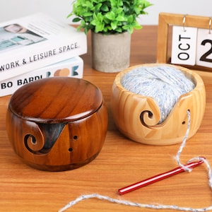 May include: Two wooden yarn bowls. One is a dark brown color with a lid, and the other is a light brown color and holds a ball of blue and white yarn. Both bowls have a swirl design and holes for the yarn to pass through. A red crochet hook is also visible.