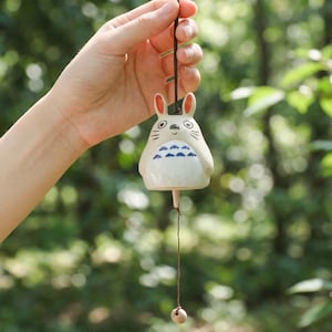 May include: A white ceramic wind chime shaped like a cartoon character with a rabbit-like face and blue markings. The wind chime is suspended by a brown string and held by a hand against a blurred green background.