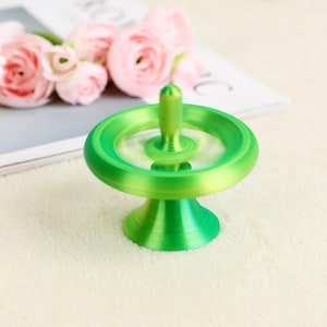 May include: A bright green spinning top with a rounded base and a central spindle. The top is in motion, creating a blur effect. The background includes pink flowers and a white surface.