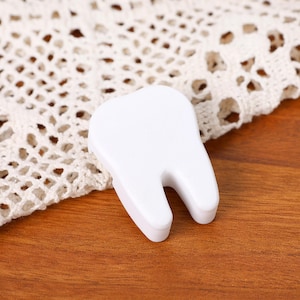 May include: A small, white, tooth-shaped object rests on a wooden surface. The tooth has a simple, clean design with a U-shaped root. A white, lacy fabric is partially visible in the background. The overall aesthetic is clean and minimalist.