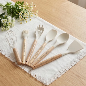 K&ouml;nnte beinhalten: Ein Set aus sechs K&uuml;chenutensilien, darunter ein Schneebesen, ein Pinsel, eine Nudelforke, ein L&ouml;ffel und ein Spatel. Jedes Utensil hat einen hellfarbigen Silikonkopf und einen Holzgriff. Der Text "Cathy's Kitchen" ist auf jedem Griff aufgedruckt.