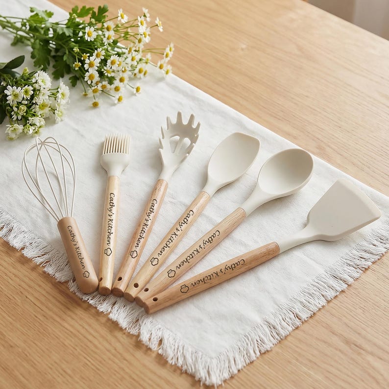 K&ouml;nnte beinhalten: Ein Set aus sechs K&uuml;chenutensilien, darunter ein Schneebesen, ein Pinsel, eine Nudelforke, ein L&ouml;ffel und ein Spatel. Jedes Utensil hat einen hellfarbigen Silikonkopf und einen Holzgriff. Der Text "Cathy's Kitchen" ist auf jedem Griff aufgedruckt.