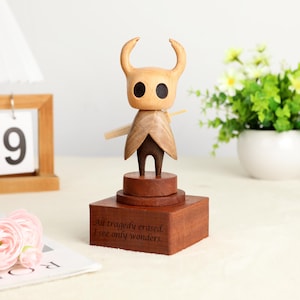 May include: A wooden figurine of a character with a horned head, large eyes, and a cape. The figurine is on a tiered base with the engraved text "All tragedy erased. I see only wonders." The figure is made of light and dark brown wood.
