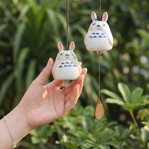 May include: Two ceramic wind chimes shaped like a cartoon character with white bodies, pink ears, and blue accents. Each chime hangs from a brown string with a wooden or ceramic clapper. The background is blurred green foliage.