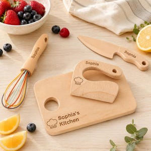 Könnte beinhalten: Ein Küchenset aus Holz mit einem Schneidebrett, das mit 'Sophia's Kitchen' und einem Kochmützen-Symbol graviert ist, einem Schneebesen mit 'Sophia'-Gravur am Griff, einem Holzmesser mit 'Sophia'-Schriftzug und einem Holzschneider mit 'Sophia'.