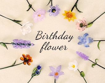 May include: A collection of glass flowers in various colors, including purple, blue, pink, yellow, and orange. The flowers are arranged in a circle around the words "Birthday flower".