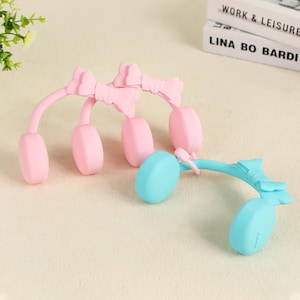 May include: Three pairs of pink and blue headphones with a bow design. The headphones have a curved band and oval earcups. The bow is at the top of the band. The headphones are made of a soft, matte material.