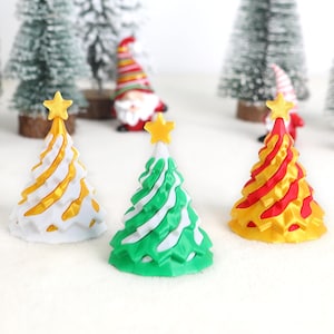 May include: Three miniature Christmas trees in white, green, and yellow with star toppers. The trees have a swirled design and are set against a backdrop of snowy miniature evergreen trees and festive gnomes.