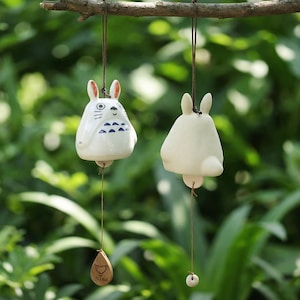 May include: Two ceramic wind chimes hanging from a tree branch. One is white with blue details and a cartoon face. The other is a plain white bell shape. Both have a wooden charm with the name "Nina".