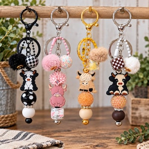 May include: Four decorative keychains hanging in a row, each featuring a highland cow charm, colorful beads, and a fluffy pom-pom. The keychains include black, pink, gold, and silver metal hardware with various patterned and solid-colored beads.