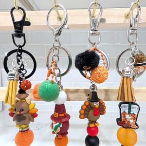 May include: Four fall-themed keychains with various charms. One features a turkey, another a gnome, a third a turkey with a hat, and the last a pumpkin spice latte. Each keychain has a tassel and decorative beads in fall colors.