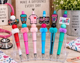 May include: Six retractable pens arranged in a clear plastic stand. Each pen features a unique topper, including a stethoscope, coffee cup, scrub top, and nurse-themed signs. The pens have colorful barrels, some glittery or metallic, and are accented with fluffy pom-poms and beads.