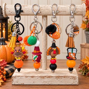 May include: Four decorative keychains hanging on a wooden stand. The first features a turkey, yellow tassel, and orange glitter bead. The second has a gnome in a hat and green pom-pom. The third shows a turkey and black pom-pom. The fourth has a coffee cup charm and brown tassel.