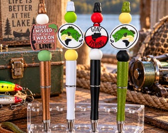 May include: Four pens with fishing-themed toppers stand in a clear acrylic holder. The first pen is copper with a sign reading 'SORRY I MISSED YOUR CALL I WAS ON THE OTHER LINE'. The others feature fish graphics and a 'GONE FISHING' bobber design.