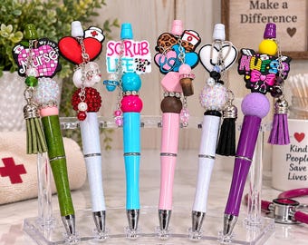 May include: Six decorative pens arranged on a clear stand, each featuring nurse-themed charms like hearts, stethoscopes, and text reading Nurse or Scrub Life. The pens have colorful barrels, glittery beads, and hanging tassels in various shades.